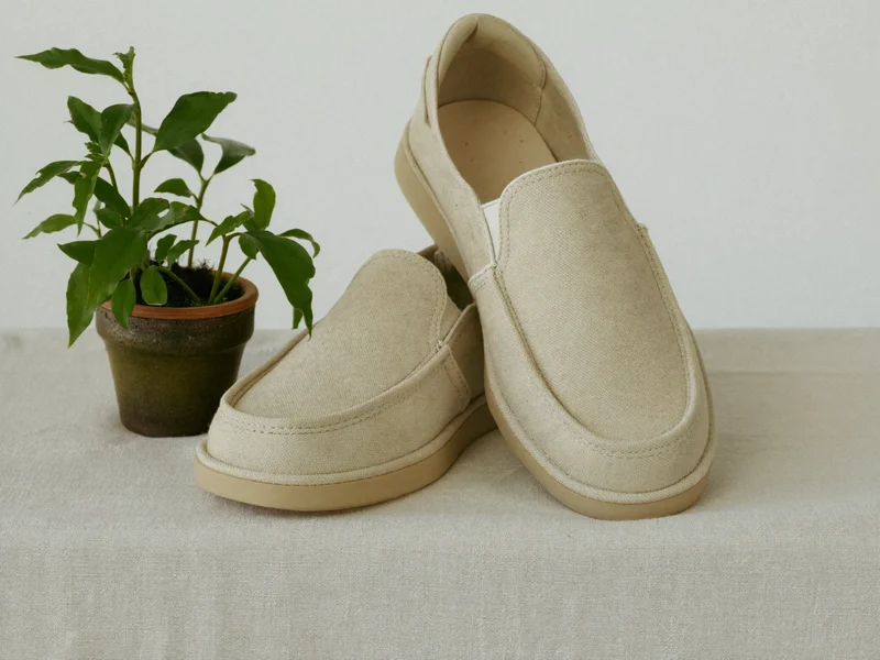 Sandfarbene Canvas-Slip-ons auf Leinentuch neben einer Zimmerpflanze, illustriert